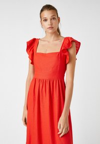 Vestido de línea roja con escote cuadrado y mangas cortas con volantes; cuenta con un cuerpo ajustado y una falda fluida, mostrando un acabado texturizado.