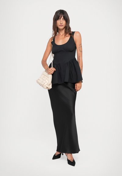 Black peplum top with a scoop neckline, paired with a long black satin skirt. Model holds a cream woven handbag and wears black heels.