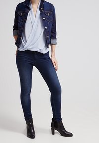 Giacca in denim con chiusura a bottoni e due tasche frontali, indossata sopra una blusa azzurro chiaro. Jeans skinny blu scuro e stivali neri con tacco alla caviglia.