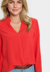 Camicia rossa con scollo a V, dettagli con colletto arricciato, maniche lunghe e tessuto morbido e leggero. Abbinata a una cintura marrone e jeans di denim.