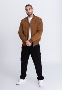 Bruine bomberjack met geribbelde manchetten en kraag, voorzien van een ritszak op de mouw. Gecombineerd met zwarte cargobroek en witte sneakers.