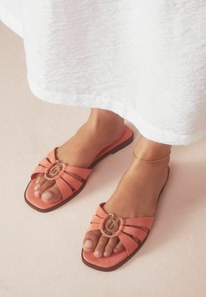 Füße, die korallrosa offene flache Sandalen mit goldfarbenem Schnallendetail tragen, kombiniert mit einem weißen Kleid und einem filigranen Fußkettchen.