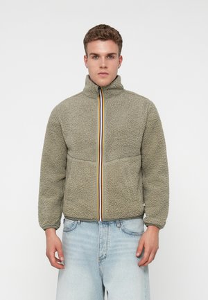 Joven que lleva una chaqueta de forro polar gris claro con collar alto y cremallera a rayas de colores, combinado con unos jeans azul claro, de pie frente a un fondo blanco.