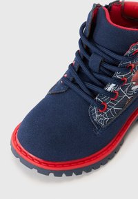 Botte bleue marine avec des accents rouges, empeigne en tissu présentant un motif de toile d'araignée, semelle en caoutchouc, lacets ronds, et un col rembourré pour le confort.