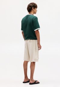 T-shirt en coton vert avec des finitions blanches, associé à un short beige avec des poches. Le mannequin porte des sandales noires. Design minimaliste, coupe décontractée.