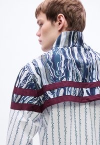 Πουκάμισο με σχέδιο, μπλε και λευκές ρίγες, μπορντό λεπτομέρειες και αφηρημένα σχέδια. Διαθέτει ψηλό γιακά και είναι φτιαγμένο από ελαφρύ ύφασμα.