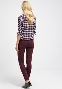 Blond kvinna som står med ryggen mot kameran, iklädd en uppvikt rutiga skjorta, burgundy tighta byxor och svarta skor mot en vit bakgrund.