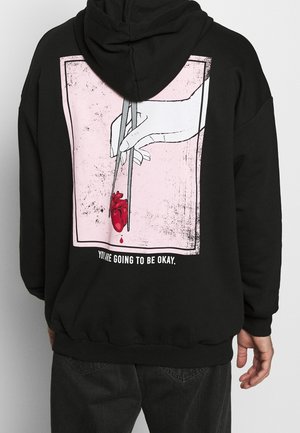 Hand, der Essstäbchen hält und ein blutendes rotes anatomisches Herz greift, mit dem Text „Du wirst in Ordnung sein“ auf der Rückseite eines schwarzen Kapuzenpullovers.