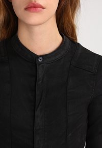 Czarna koszula z denimu z okrągłym kołnierzykiem, zapięciem na guziki i subtelną teksturą. Posiada pionowe szwy na froncie.