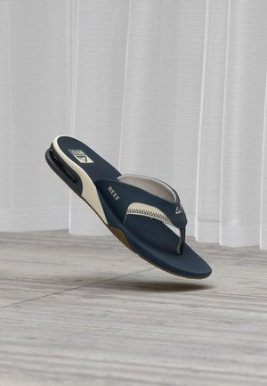 Sandale tongs Reef bleu marine et blanche avec semelle matelassée et poche d’air au talon, montrée flottant au-dessus d’un sol gris avec des rideaux blancs translucides.