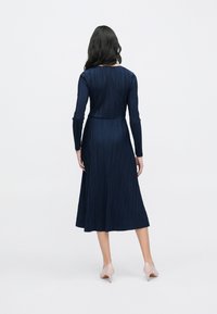 Tengerészkék, pliszírozott ruha hosszú ujjakkal és testhezálló derékkal, középmellfesztávolságig érő hosszban, bézs magas sarkú cipővel párosítva.