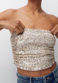 Top sans manches en sequins dorés et argentés avec une texture froncée, un design ajusté et un matériau extensible. Associez-le à un bas en denim avec une taille visible.