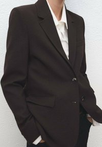 Brązowa, dopasowana marynarka z usztywnionymi ramionami, dwoma przednimi kieszeniami i dwoma guzikami. Noszona na białej koszuli z kołnierzykiem. Gładka struktura tkaniny.