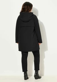 Cappotto impermeabile nero con cappuccio, caratterizzato da un taglio comodo, maniche lunghe e lunghezza fino a metà coscia. Abbinato a pantaloni neri con texture e stivaletti con tacco.