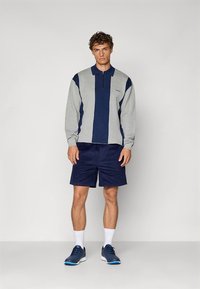 Polo de mangas compridas, com blocos de cor cinza e azul-marinho e gola com fecho de correr, combinado com calções de bombazina azul-marinho e ténis desportivos azuis com detalhes em branco.