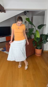 Femme portant un chemisier orange et une jupe blanche, regardant vers le bas, soulevant légèrement sa jupe tout en se tenant sur un sol en bois à côté d'une grande plante en pot.