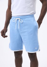 Pantalones cortos de algodón azul claro con cordón blanco, bolsillos laterales y ribete blanco. Presenta un pequeño logo en la parte inferior de la pierna.