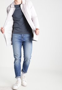 Valkoinen kevyt takki nappeilla, tummansininen T-paita, vaaleansiniset slim-fit farkut ja valkoiset tennarit, esittelee rentoa istuvuutta ja epämuodollista tyyliä.