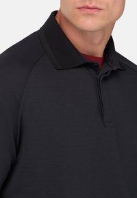 Svart polo skjorta med texturerad tyg, klassisk krage och knappslå. Under finns en röd rundhalsad t-shirt.