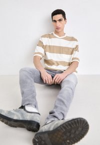 Randig kortärmad skjorta i beige och vitt med bröstficka, matchad med ljusgrå jeans och grå skor, mot en neutral bakgrund.