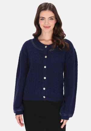 Donna sorridente con capelli castani ondulati che indossa un cardigan blu scuro a fantasia con bottoni e una gonna nera su uno sfondo uniforme.