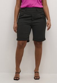 Shorts noirs avec des revers, présentant un devant plat, des plis et un tissu lisse. Portés avec un haut rose et des sandales noires à lanières.