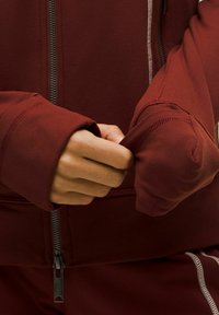 Giacca sportiva bordeaux con chiusura a zip, caratterizzata da un design aderente e polsini a costine. Texture liscia con rifiniture in grigio.