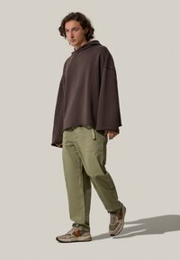 Felpa oversize marrone, pantaloni cargo verdi con tasche e sneakers marroni con dettagli arancioni. Lo sfondo neutro mette in risalto i dettagli degli abiti.