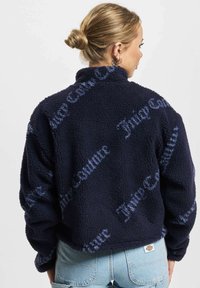 Ženska z blond lasmi v koškem pričesku nosi mornarsko modro puhasto jakno z napisom "Juicy Couture" in svetlobluje džins hlače, prikazana s hrbta.