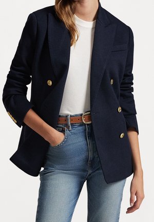 Person iført en marineblå dobbeltbrystet blazer med guldknapper, hvid t-shirt, brun bælte og lyseblå jeans, med den ene hånd i lommen.