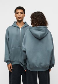 Φούτερ με φερμουάρ σε τιρκουάζ χρώμα, με ευρύχωρη μπροστινή τσέπη, κουκούλα με κορδόνι και φαρδιά εφαρμογή. Μαλακό ύφασμα με λεία υφή και ριμπ μανσέτες.