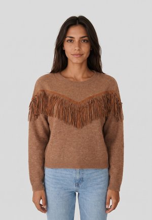 Donna che indossa un maglione marrone con frange e dettagli borchiati sul petto, abbinato a jeans azzurri chiari, in piedi davanti a uno sfondo uniforme.