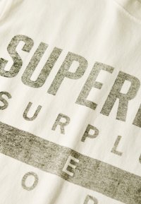 Nærbillede af falmet sort tekst "SUPER SURPLUS" trykt på off-white stof med synlig tekstur.