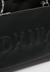 Sac à main en cuir noir avec logo embossé, ferrures argentées, design structuré et fermeture éclair supérieure. Texture lisse sans motifs visibles.