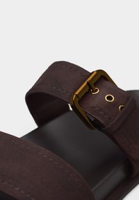 Κοντινό πλάνο καφέ σουέτ λουριού σανδαλιού με χρυσή πόρπη και μαύρη σόλα σε λευκό φόντο.