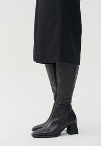 Botas de cuero negras hasta la rodilla con punta afilada, tacón ancho y textura brillante. Cuentan con un zipper lateral para el cierre.