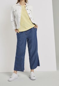 Chaqueta de mezclilla blanca, camiseta a rayas amarillas y blancas, pantalones anchos azules y zapatillas blancas; ajuste relajado y estilo casual.