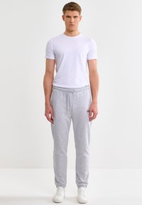 Giovane uomo in piedi, indossa una maglietta bianca a maniche corte, pantaloni della tuta grigio chiaro con coulisse e scarpe da ginnastica bianche su uno sfondo bianco semplice.