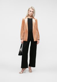 Femme aux cheveux blonds portant un blazer en velours beige, un haut noir et un pantalon noir, tenant un sac à main noir et portant des sandales à talons blancs.