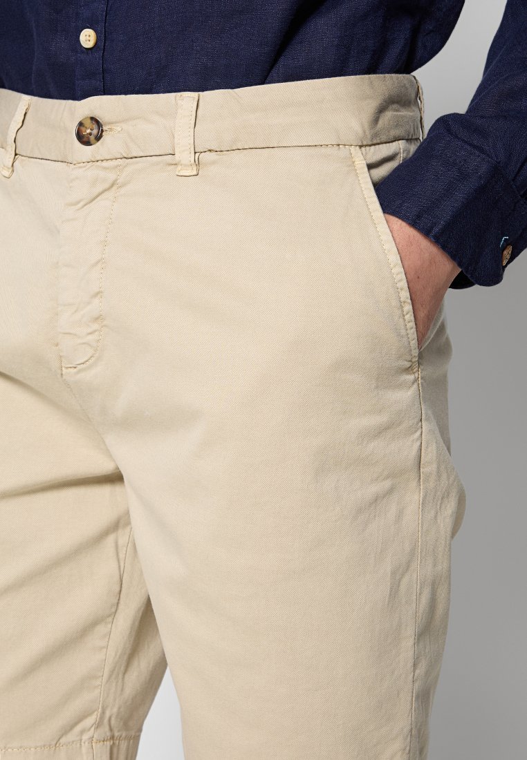 Jasno beżowe bawełniane chinosy o klasycznym kroju, z bocznymi kieszeniami, płaskim przodem i zapięciem na guzik. Teksturowana tkanina o gładkim wykończeniu.