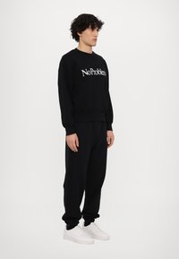 Sudadera negra con el texto blanco "NoProblem", combinada con pantalones de chándal negros y zapatillas blancas. Estilo casual, corte relajado con puños acanalados.