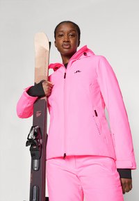 Chaqueta de esquí rosa con pantalones a juego, que presenta un diseño ajustado, frente con cremallera y acentos en negro. Esquís sostenidos con una mano.