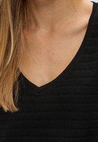 Ženský krk a klíční kost s lehce světlou pletí a několika pihami, oblečená v černém ribbed topu s V-výstřihem, s hnědými rovnými vlasy spadajícími na levou stranu.