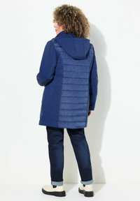 Giacca da esterno blu navy con pannello posteriore trapuntato, cappuccio e maniche lunghe. Si abbina a jeans in denim scuro e stivaletti beige alla caviglia.