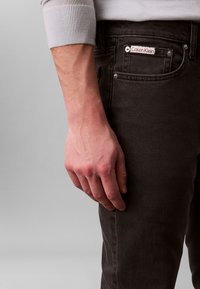 Black denim jeans with a Calvin Klein label; close-up of a hand resting on the pocket, showcasing texture and stitching details.