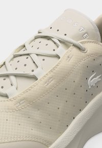 Μπεζ αθλητικό παπούτσι Lacoste με διάτρητο επάνω μέρος, λευκά κορδόνια, χαραγμένο όνομα μάρκας στη γλώσσα και μικρό λογότυπο κροκόδειλου στο πλάι.