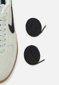 Light gray suede sneaker with a black swoosh, gray laces, and a brown rubber sole, accompanied by two coiled black shoelaces.