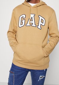 Sweat à capuche beige avec le logo "GAP" en blanc et bleu marine, doté d'une capuche à cordon de serrage et d'une poche kangourou. Porté avec un jean bleu déchiré.
