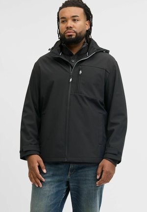 Homme avec barbe et dreadlocks portant une veste à capuche noire par-dessus un pull boutonné noir et un jean bleu, se tenant devant un fond blanc.