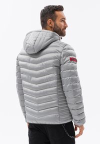 Grå vadderad jacka med huva, med ett chevron-quiltat mönster och en livlig logotyp patch på ärmen. Slät, glansig tygtextur.